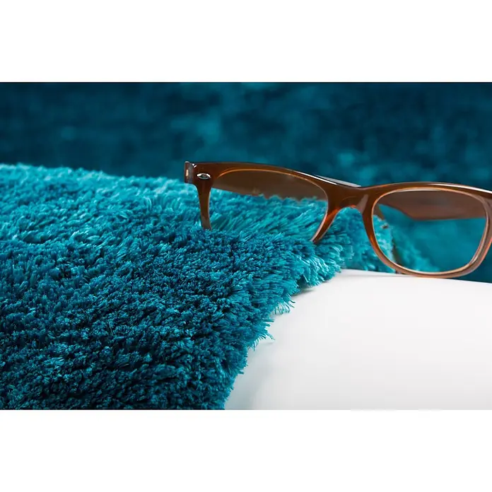 Kayoom Hochflorteppich Dolphin 500 - Blau, 290 x 200 cm, 100 % Polyester - Braune Brille ruht auf einem plüschigen, türkisfarbenen Hochflorteppich.