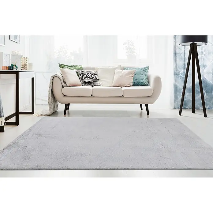 Kayoom Fellteppich Emotion 525 - Silber, 150 x 80 cm, 100 % Polyester - Ein hellgrauer, flauschiger rechteckiger Teppich liegt auf einem Holzboden in einem Wohnzimmer vor einem Sofa.