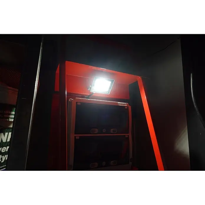 Baustromverteiler Doktorvolt 1x CEE 32A, 2x CEE 16A, 4x SCHUKO, 1x LED-Strahler, mit Rollen - Anzahl CEE-Steckdosen: 3 Stk., Anzahl Steckdosen Schuko: 4 Stk., IP44 - Helle LED-Flutlichtleuchte an rotem Gestell, beleuchtet dunkle Elektrogeräte darunter.