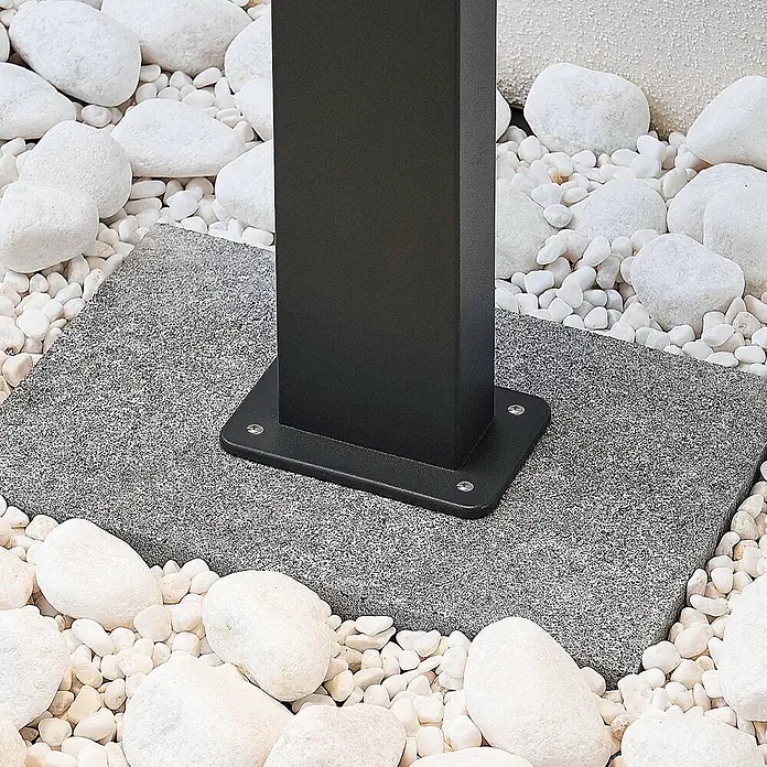 Lindby LED Wegeleuchte in anthrazit, weiß, Gartenlampe -  - Schwarzer Vierkantpfosten mit robuster Bodenplatte auf grauer Granitplatte, umgeben von weißen Zierkieseln.