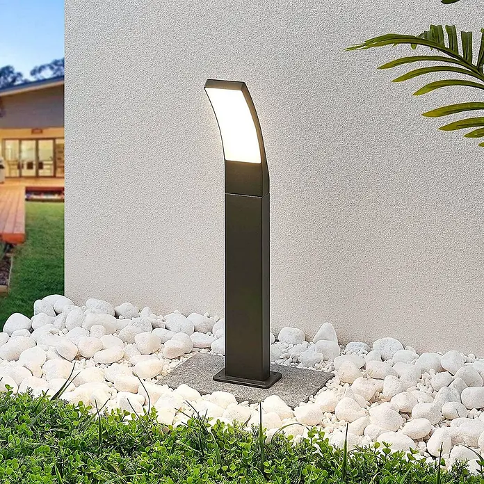 Lindby LED Wegeleuchte in anthrazit, weiß, Gartenlampe -  - Dunkelgraue Außenstandleuchte mit gebogener, beleuchteter Oberseite, auf grauem Sockel zwischen weißen Kieselsteinen und Pflanzen im Garten.
