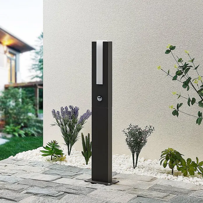 Lucande LED Wegeleuchte in graphit, weiß, mit Bewegungsmelder, Gartenlampe -  - Eine hohe schwarze Außenwegleuchte mit weißem Diffusorstreifen und Bewegungssensor steht auf einem Steinweg neben Gartenpflanzen.