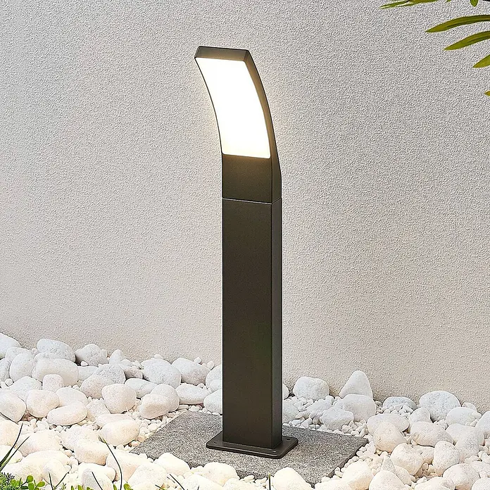 Lindby LED Wegeleuchte in anthrazit, weiß, Gartenlampe -  - Moderne anthrazitfarbene Pollerleuchte mit gekrümmter, beleuchteter Oberseite, in einem Kiesbeet platziert.