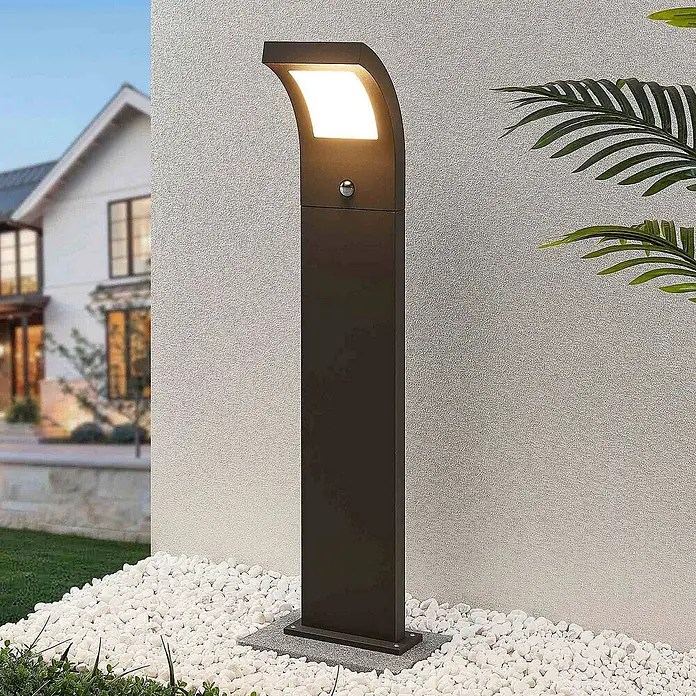 Arcchio Außenlaterne Advik -  - Dunkle Außenleuchte mit gebogenem Kopf und Sensor, beleuchtet weiße Kieselsteine vor einer Hauswand.