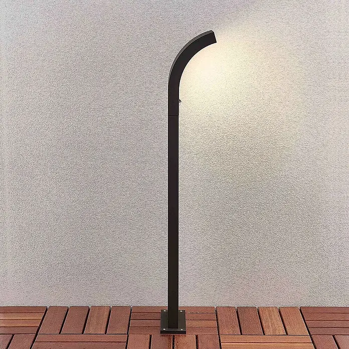 Arcchio Außenlaterne Advik -  - Eine schwarze, moderne, gebogene Außenwegleuchte steht auf einer Holzterrasse und beleuchtet eine helle Strukturwand.