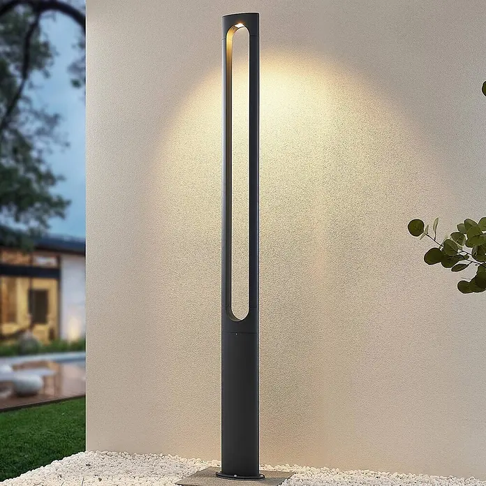 Lucande LED Gartenlaterne für draußen in dunkelgrau, Mastleuchte -  - Hohe, dunkelgraue Außenpollerleuchte mit bogenförmigem Ausschnitt, die warmes Licht auf eine Wand in einer Gartenumgebung abgibt.