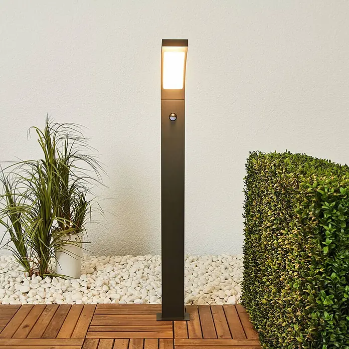 Lucande Außenlaterne Juvia -  - Hohe, dunkle Pollerleuchte für den Außenbereich mit beleuchteter Oberseite und integriertem Sensor, auf Holzterrasse platziert.