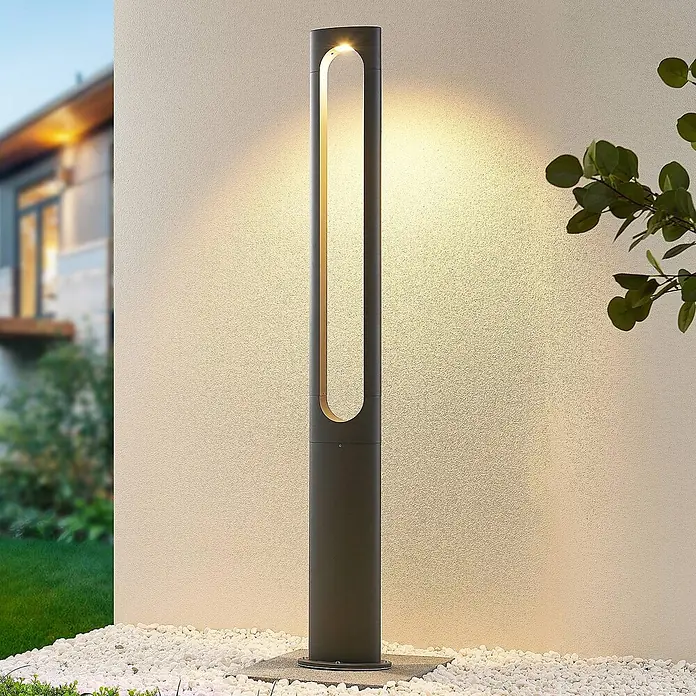 Lucande LED Gartenlaterne für draußen in dunkelgrau, Mastleuchte -  - Dunkelgraue Wegeleuchte beleuchtet eine strukturierte Wand und weiße Kiesel im Garten.