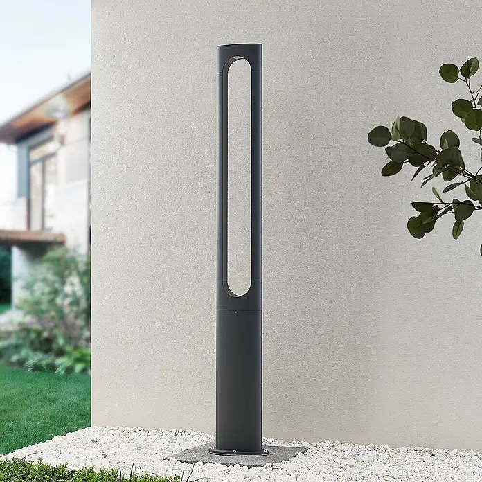 Lucande LED Gartenlaterne für draußen in dunkelgrau, Mastleuchte -  - Eine hohe, dunkelgraue moderne Pollerleuchte für den Außenbereich mit ovalem Ausschnitt, auf weißen Kieselsteinen an einer Wand.