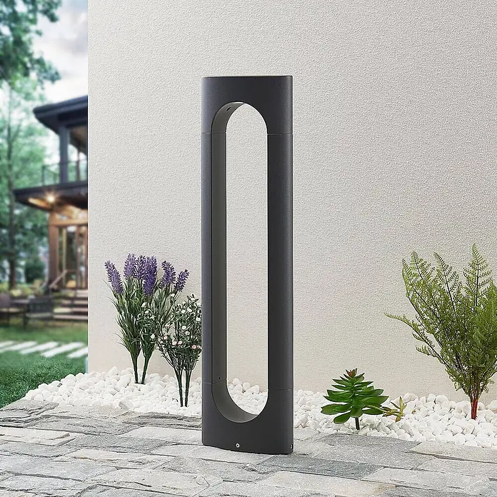 Lucande Außenlaterne Fenti -  - Dunkelgraue, moderne Pollerleuchte mit ovalem Ausschnitt, beleuchtet einen gepflasterten Gartenweg neben einem Haus und Pflanzen.