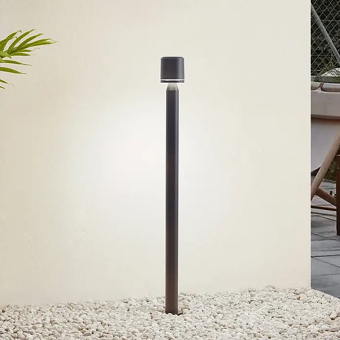 Lindby Außenlaterne Amila -  - Dunkle Metall-Pollerleuchte für den Außenbereich beleuchtet eine cremefarbene Wand, stehend auf weißen Kieselsteinen.