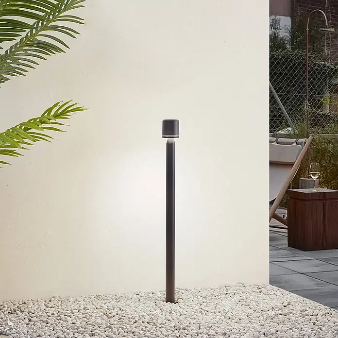 Lindby Außenlaterne Amila -  - Dunkle Pollerleuchte erhellt einen Kiesweg in einer Außenanlage.
