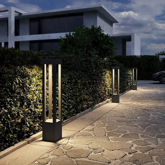 Arcchio LED Wegeleuchte in dunkelgrau, Gartenlampe -  - Moderne dunkle Außenpollerleuchte mit beleuchteten Streifen, die warmes Licht auf eine Hecke und einen Steinweg wirft.