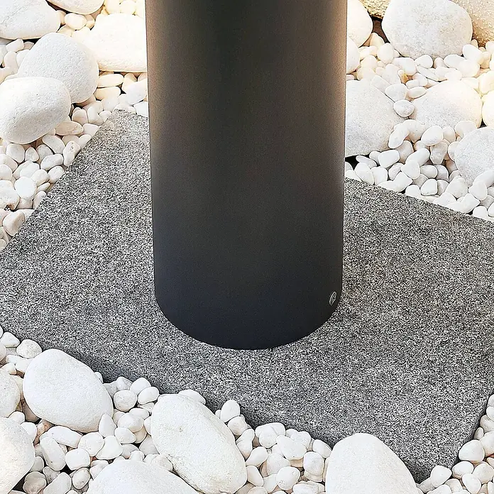 Lucande LED Wegeleuchte, Pollerleuchte Dunkelgrau, Außenleuchte 65 cm -  - Schwarze zylindrische Leuchte auf einer grauen Steinplatte, umgeben von weißen Zierkieseln.