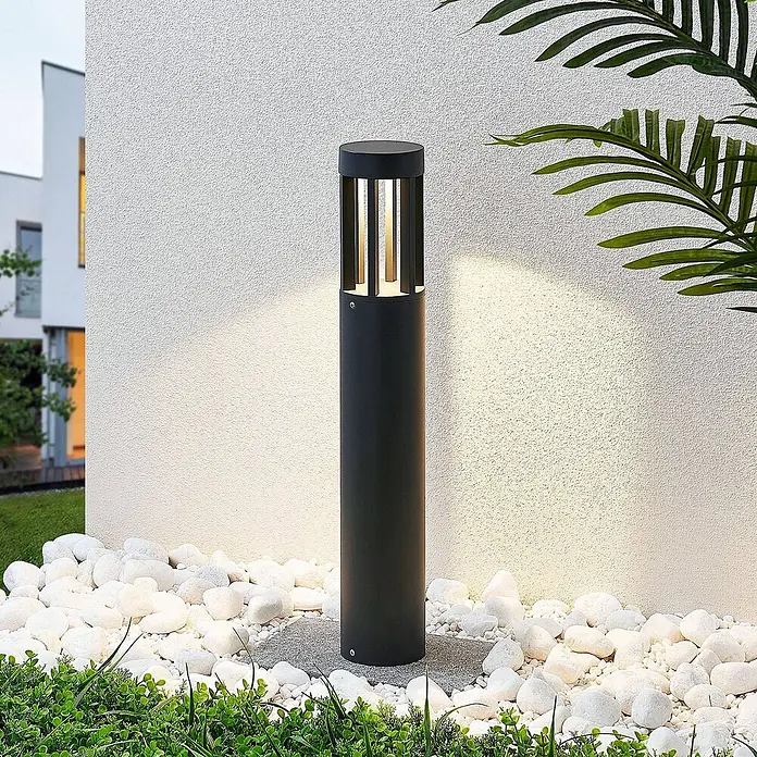 Lucande LED Wegeleuchte, Pollerleuchte Dunkelgrau, Außenleuchte 65 cm -  - Schwarze zylindrische Wegeleuchte beleuchtet eine strukturierte weiße Wand und weiße Kieselsteine im Garten.