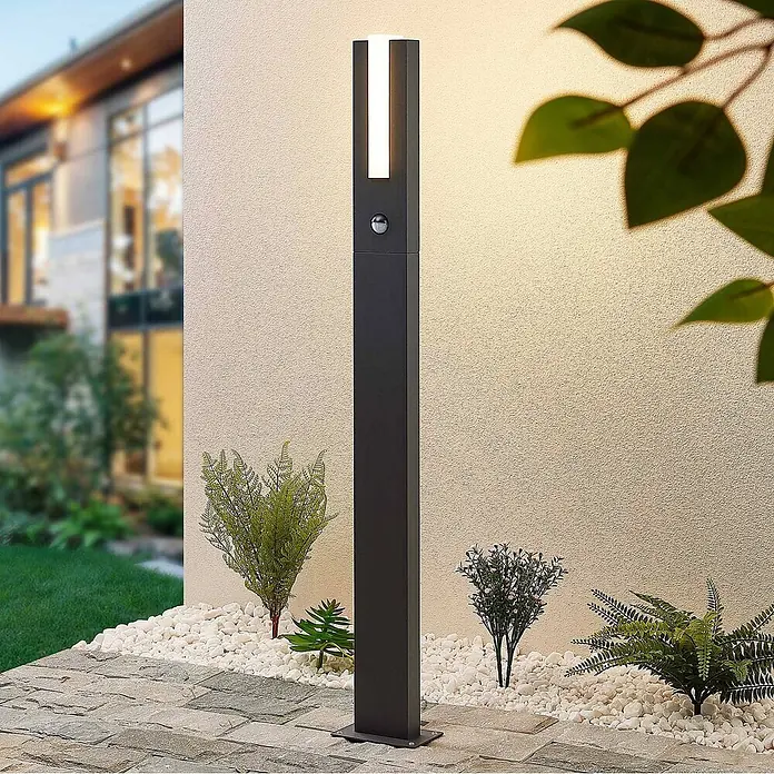 Lucande Außenlaterne Virgalia -  - Dunkelgraue, rechteckige Außenwegleuchte mit vertikalem LED-Streifen und Bewegungsmelder, beleuchtet eine Hauswand, umgeben von weißen Kieseln und Pflanzen.