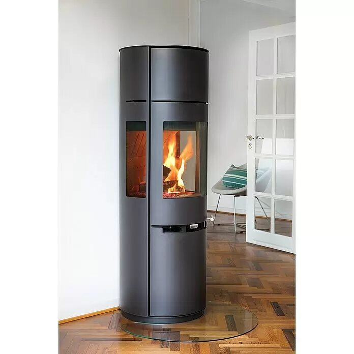 Aduro Kaminofen 9-7 - 8 kW, Raumheizvermögen: 334 m³, Material Verkleidung: Stahl, Schwarz - Dunkelgrauer zylindrischer Holzofen mit brennendem Feuer, auf Glasplatte auf Holzboden im Raum.