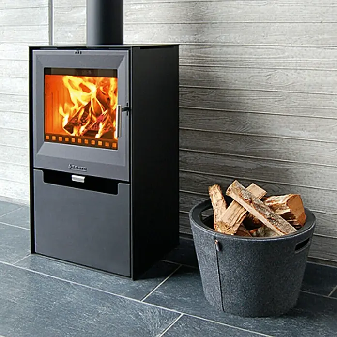 Aduro Kaminofen 14 - 6,5 kW, Raumheizvermögen: 334 m³, Material Verkleidung: Stahl, Schwarz - Ein Holzkaminofen mit brennendem Feuer und daneben ein Korb voller Holzscheite, die für Wärme und Gemütlichkeit sorgen.