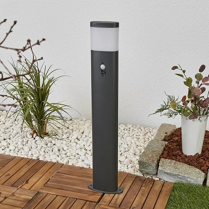 Lindby Außenlaterne Obri -  - Dunkelgraue Wegeleuchte mit Bewegungssensor, weißem Diffusor, auf Holzterrasse neben weißen Kieselsteinen installiert.
