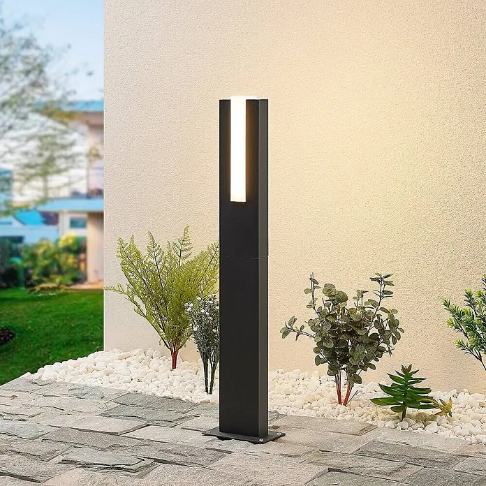 Lucande LED Wegeleuchte in anthrazit, weiß, Gartenlampe -  - Eine schwarze, rechteckige Außenleuchte mit vertikalem Leuchtstreifen steht auf einer Terrasse neben weißen Kieselsteinen und Pflanzen und beleuchtet eine Wand.