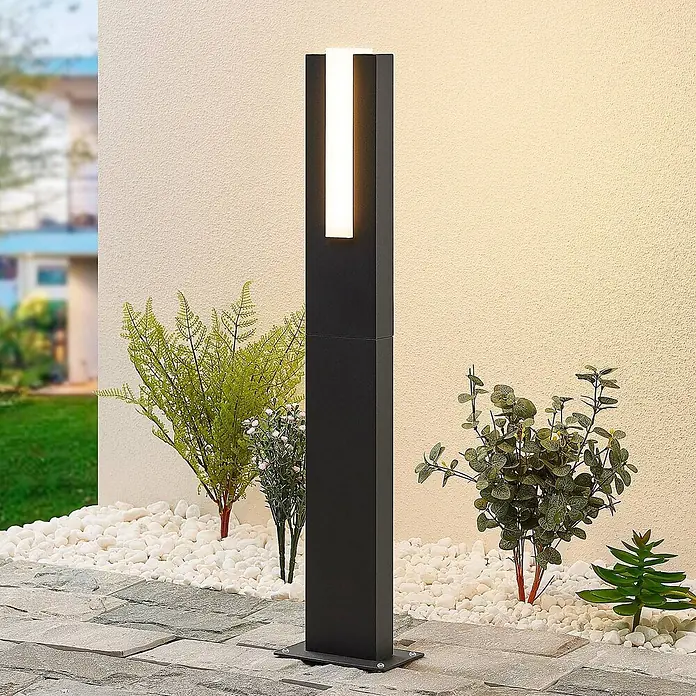 Lucande LED Wegeleuchte in anthrazit, weiß, Gartenlampe -  - Schwarze rechteckige Außenleuchte mit vertikal beleuchtetem Abschnitt, stehend zwischen weißen Kieselsteinen und Pflanzen an einer Wand.