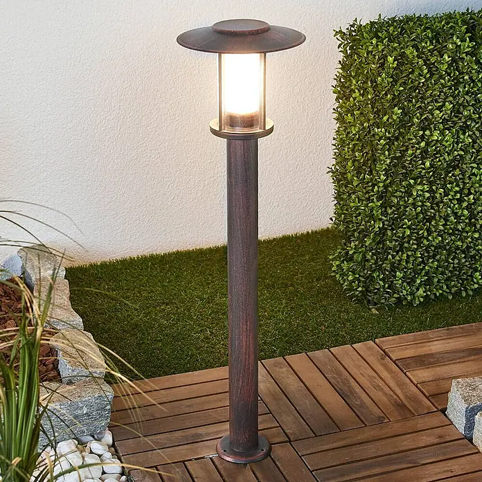 Lindby LED Wegeleuchte in rost, transparent, Gartenlampe -  - Eine braun-bronzene Pollerleuchte mit zylindrischem Diffusor beleuchtet einen Gartenweg mit Holzplatten.