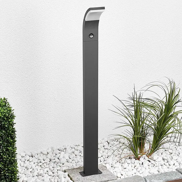 Lucande Außenlaterne Timm -  - Anthrazitfarbene Wegeleuchte mit Bewegungsmelder, in einem Garten mit weißen Kieselsteinen und Pflanzen.