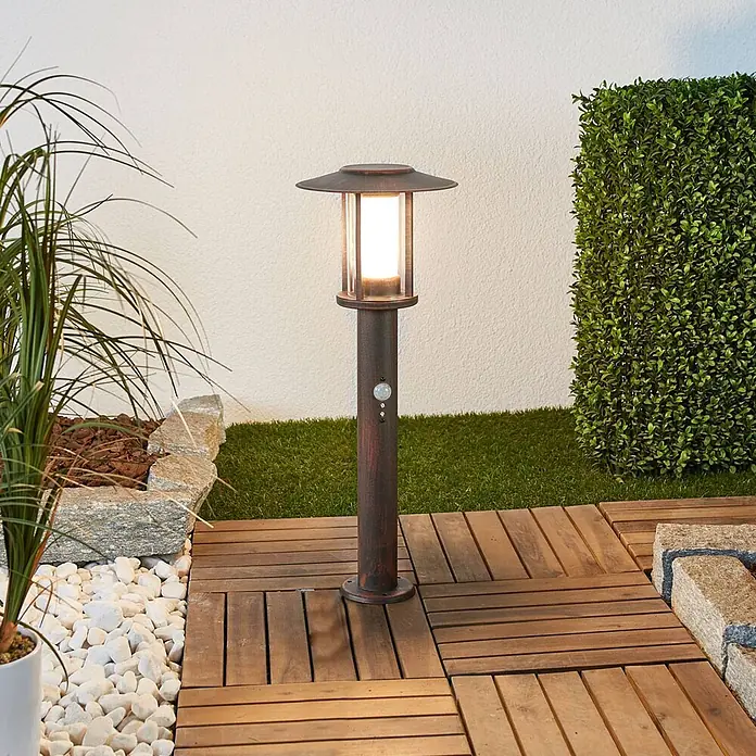Lindby Außensockelleuchte Pavlos -  - Dunkelbraune Außenwegleuchte mit Bewegungsmelder, beleuchtet einen Gartenweg auf Holzfliesen.