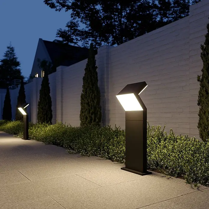 Arcchio LED Sockelleuchte in dunkelgrau, Wegeleuchten außen -  - Zwei schwarze Wegeleuchten mit heller LED-Beleuchtung säumen nachts einen Gartenweg entlang grüner Büsche und einer weißen Ziegelmauer.