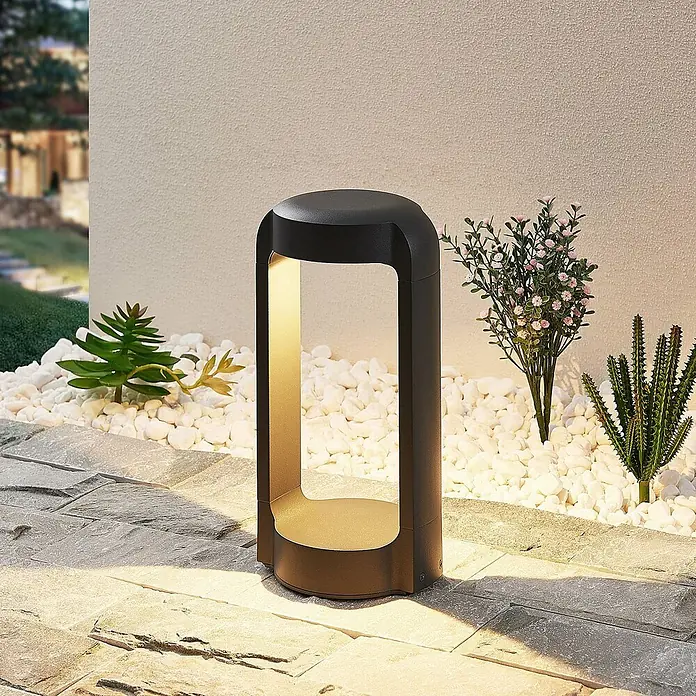 Lucande LED Sockelleuchte in anthrazit, Wegeleuchten außen -  - Dunkle LED-Wegeleuchte aus Metall mit warmem Licht, beleuchtet einen Gartenweg mit weißen Kieseln und Pflanzen.