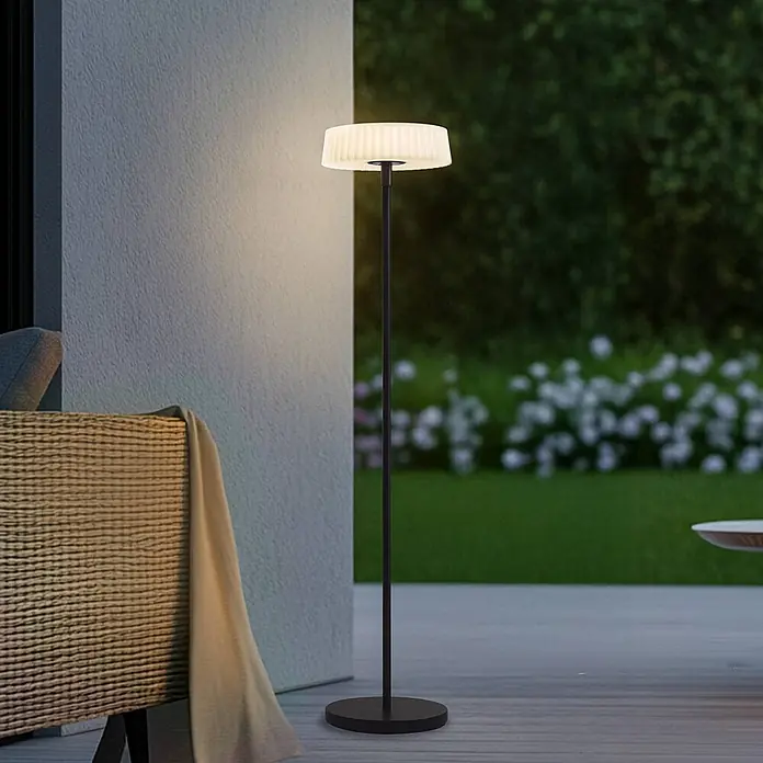 Lindby LED Solar Stehlampe Outdoor in schwarz, weiß, dimmbar Stehleuchte -  - Eine schwarze Outdoor-Stehleuchte mit einem plissierten, beleuchteten Schirm steht auf einer Terrasse und spendet stimmungsvolles Licht.