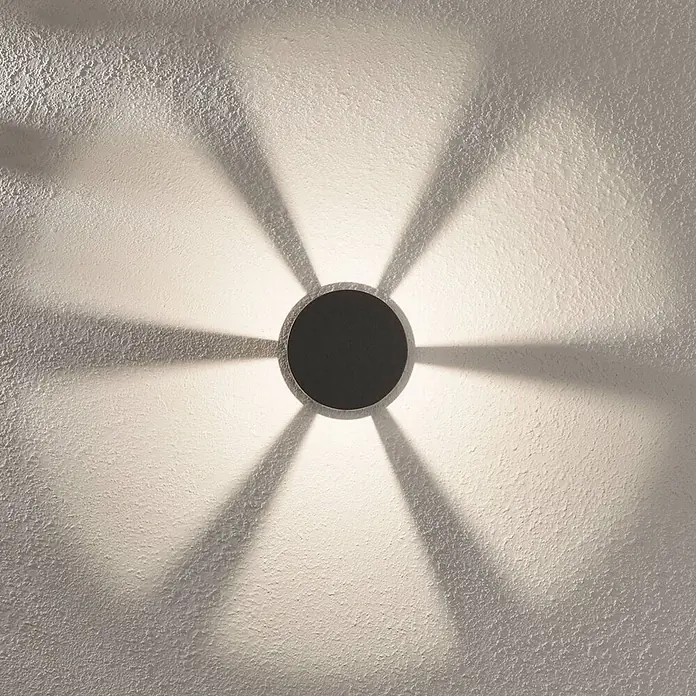 Lindby Außensockelleuchte Jordis -  - Dunkle, runde Wandleuchte, die ein sternförmiges Lichtmuster auf eine strukturierte Wand wirft.