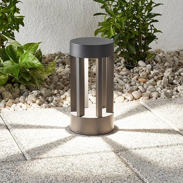 Lindby Außensockelleuchte Jordis -  - Dunkelgraue Außenpollerleuchte beleuchtet eine Terrasse mit sternförmigem Muster. Zwischen Kies und Pflanzen platziert.