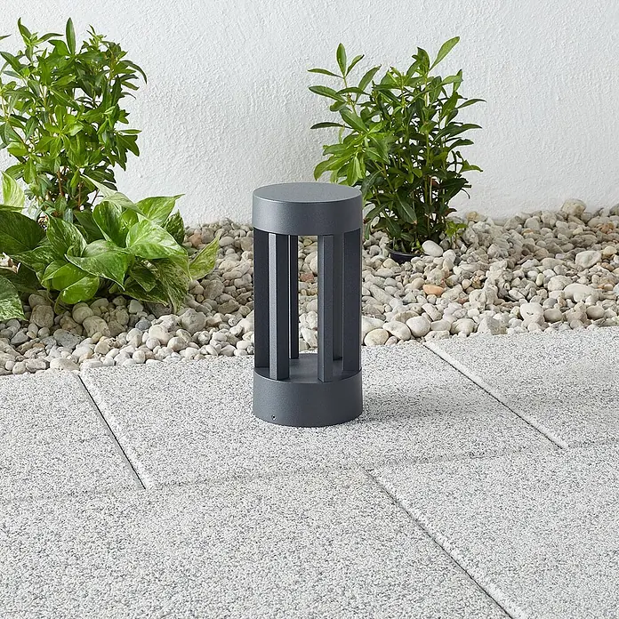 Lindby Außensockelleuchte Jordis -  - Dunkelgraue Lamellen-Pollerleuchte für den Außenbereich auf einer Terrasse neben Kieselsteinen und Pflanzen.