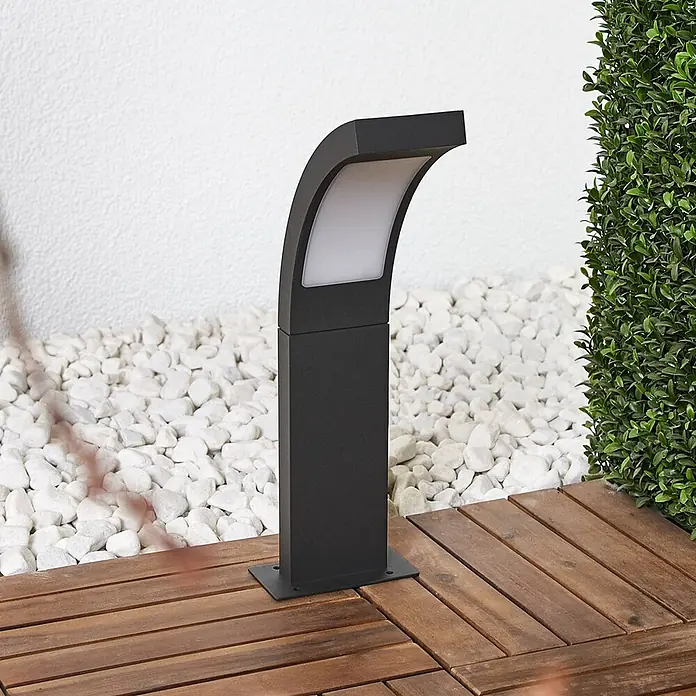 Lucande Außensockelleuchte Juvia -  - Schwarze Gartenleuchte mit gebogenem Oberteil und mattierter Scheibe, auf Holzterrasse neben weißen Ziersteinen installiert.