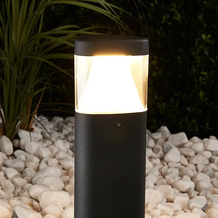 Lucande LED Sockelleuchte in anthrazit, transparent, Wegeleuchten außen -  - Schwarze Außenpollerleuchte strahlt warmweißes Licht aus, platziert zwischen weißen Kieselsteinen und Grün.