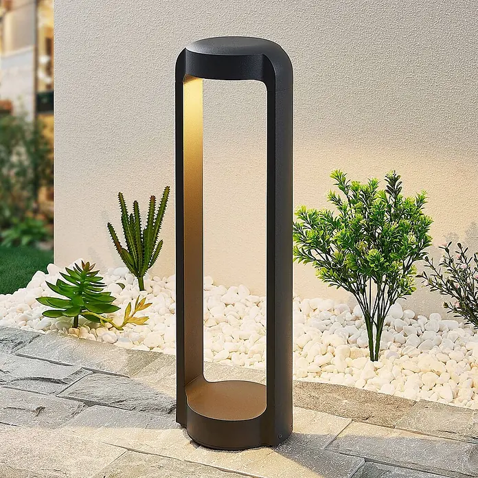 Lucande Außensockelleuchte Habsa -  - Dunkle Wegeleuchte mit goldener Innenseite beleuchtet einen Gartenweg mit weißen Steinen und Pflanzen.