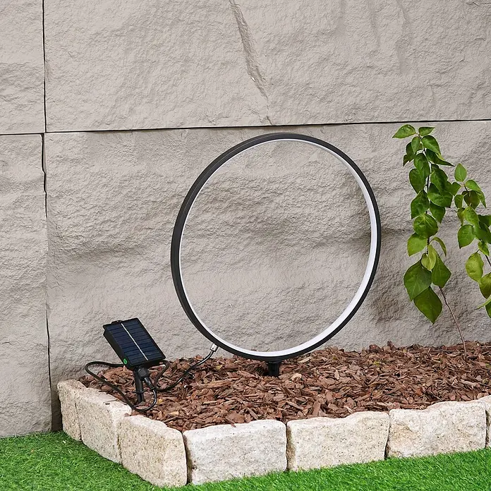 Lindby LED Solar Stehlampe Outdoor in schwarz matt, Stehleuchte Garten -  - Große schwarze LED-Ring-Gartenleuchte mit Solarpanel, im Blumenbeet mit Rindenmulch vor einer Steinmauer.