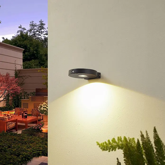 Lucande Außenwandleuchte Tavon - schwarz, schwarz, IP54 - Schwarze kreisförmige Außenwandleuchte mit Bewegungssensor, beleuchtet eine Wand in einer Gartenumgebung am Abend.