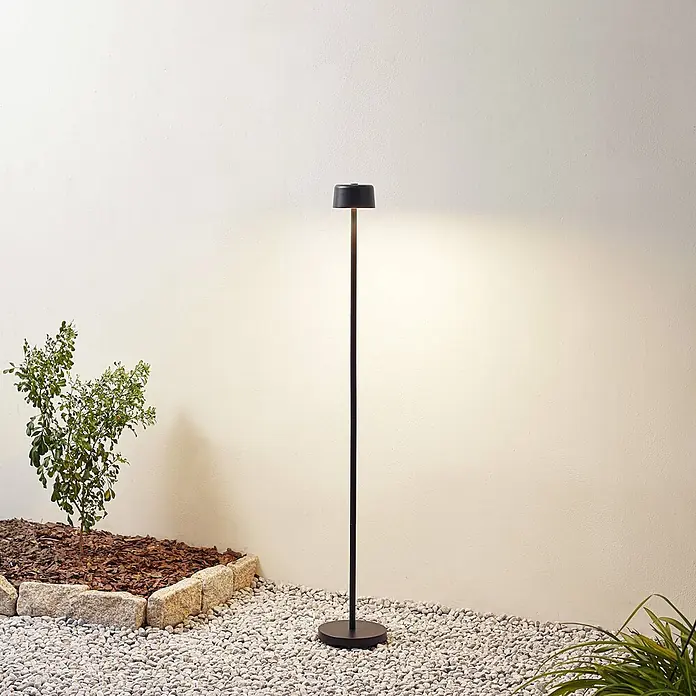 Lindby LED Stehlampe Outdoor in schwarz, dimmbar Stehleuchte Garten -  - Elegante schwarze Außenstehleuchte beleuchtet eine Außenwand, stehend auf weißen Kieselsteinen.