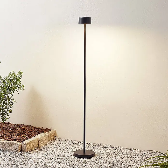 Lindby LED Stehlampe Outdoor in schwarz, dimmbar Stehleuchte Garten -  - Schlanke, schwarze Outdoor-Stehlampe mit minimalistischem Design, die eine helle Wand im Außenbereich beleuchtet.