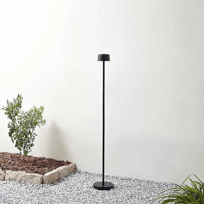 Lindby LED Stehlampe Outdoor in schwarz, dimmbar Stehleuchte Garten -  - Eine schlanke schwarze Außenstehlampe steht auf weißen Kieselsteinen in einer Gartenumgebung.