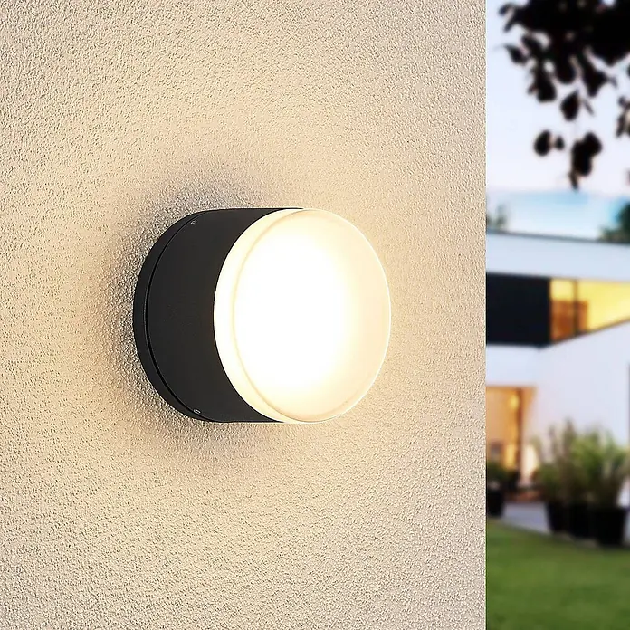 Lindby LED Wandleuchte aussen in dunkelgrau, weiß, Aussenlampe Wand -  - Runde schwarze Außenwandleuchte mit weißem Diffusor, leuchtend, an einer strukturierten Wand im Garten montiert.