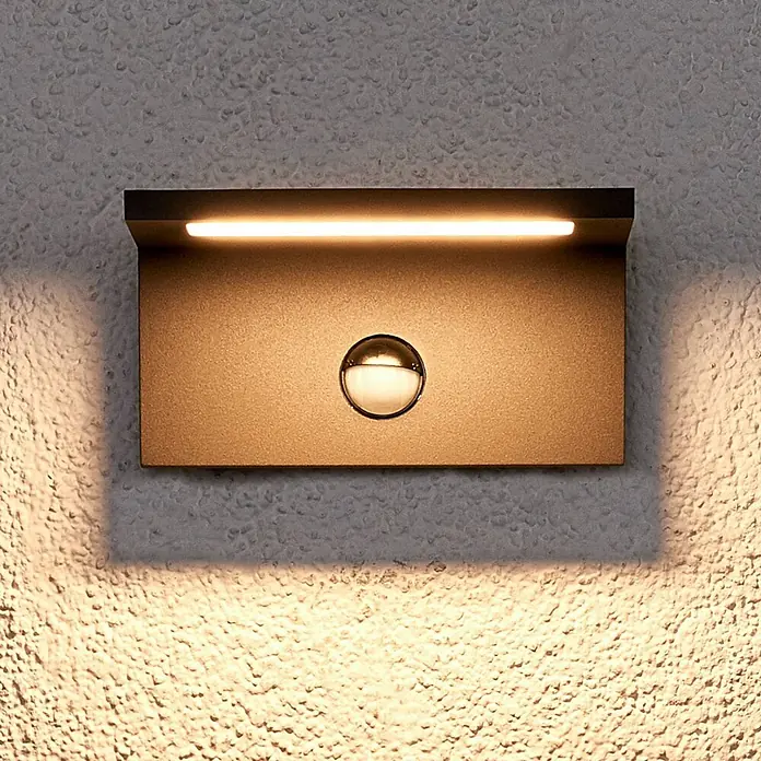 Lucande LED Wandleuchte aussen in graphit, weiß, mit Bewegungsmelder, -  - Dunkelbraune rechteckige Außenwandleuchte mit LED-Streifen oben und zentralem Bewegungssensor, an einer strukturierten Wand montiert.