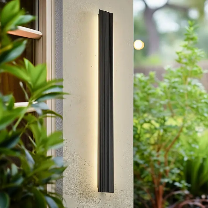 Lucande LED Wandleuchte aussen in schwarz, Aussenlampe Wand -  - Schwarze Außenwandleuchte mit vertikalen Rillen, strahlt warmes Licht nach oben und unten auf eine helle Wand, umgeben von Grün.