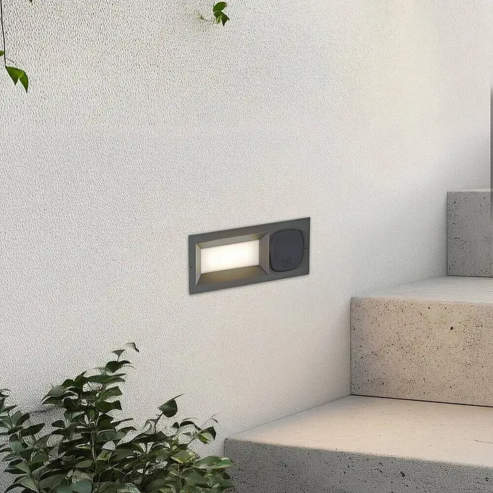 Lucande Außenwandleuchte Lachlain - schwarz, IP54 - Dunkelgraue Outdoor-Einbauleuchte an strukturierter Wand, beleuchtet Betontreppen. Mit integrierter Abdecksteckdose.