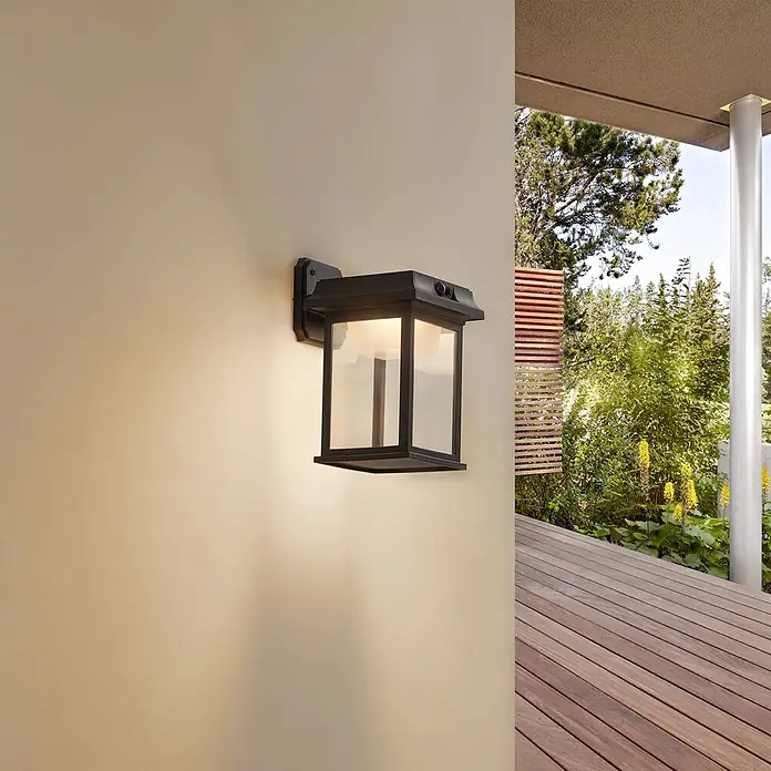 Lindby LED Solar Wandleuchte aussen in schwarz, mit Bewegungsmelder, -  - Beleuchtete schwarze Außenwandleuchte mit Klarglas, an heller Wand über Holzterrasse montiert.