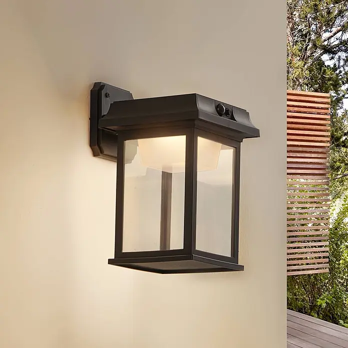 Lindby LED Solar Wandleuchte aussen in schwarz, mit Bewegungsmelder, -  - Schwarze Laternen-Wandleuchte für den Außenbereich mit klaren Glaspaneelen und Sensoren, beleuchtet an einer Wand.