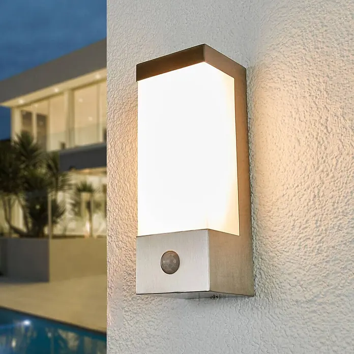 Lindby Außenwandleuchte Severina - alu / grau / zink, IP44 - Moderne rechteckige Außenwandleuchte aus gebürstetem Metall mit weißem Diffusor und Bewegungssensor, beleuchtet eine Außenwand im Abendlicht.