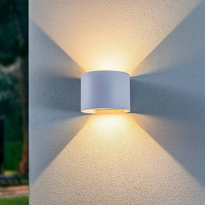 Lindby LED Wandleuchte aussen in weiß, up down Aussenlampe Wand -  - Weiße zylindrische Außenwandleuchte, die warmes Licht nach oben und unten auf eine strukturierte Wand wirft, mit Grün im Hintergrund.